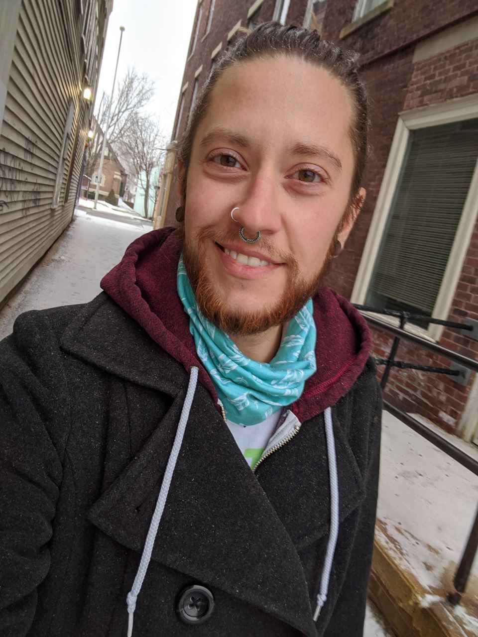 Selfie of Reid. They are white with long brown hair pulled up into a bun. They have a short beard, a nose ring and a septum ring. They are wearing a teal neckerchief, a maroon hoodie, and a grey peacoat. They are standing in an alleyway between two brick buildings and there is snow on the ground behind them.