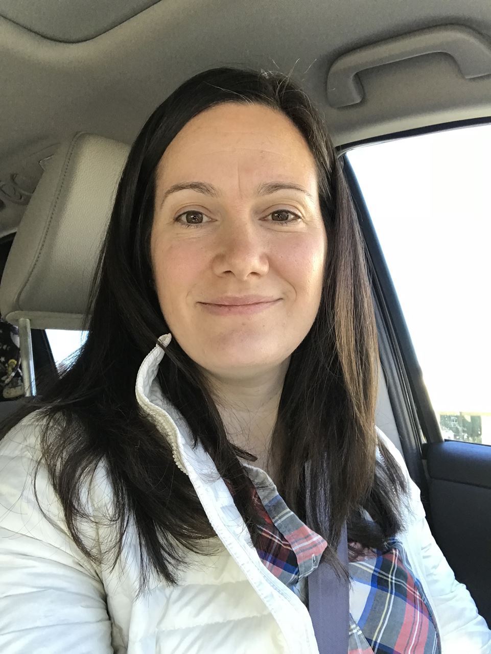 [image description White female sitting in car. She has long, straight, dark brown hair and is wearing a plaid shirt and white jacket. She is smiling at the camera.]